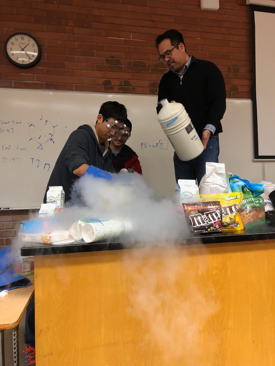 One person pouring liquid nitrogen into a container while another person stirs and a third person watches